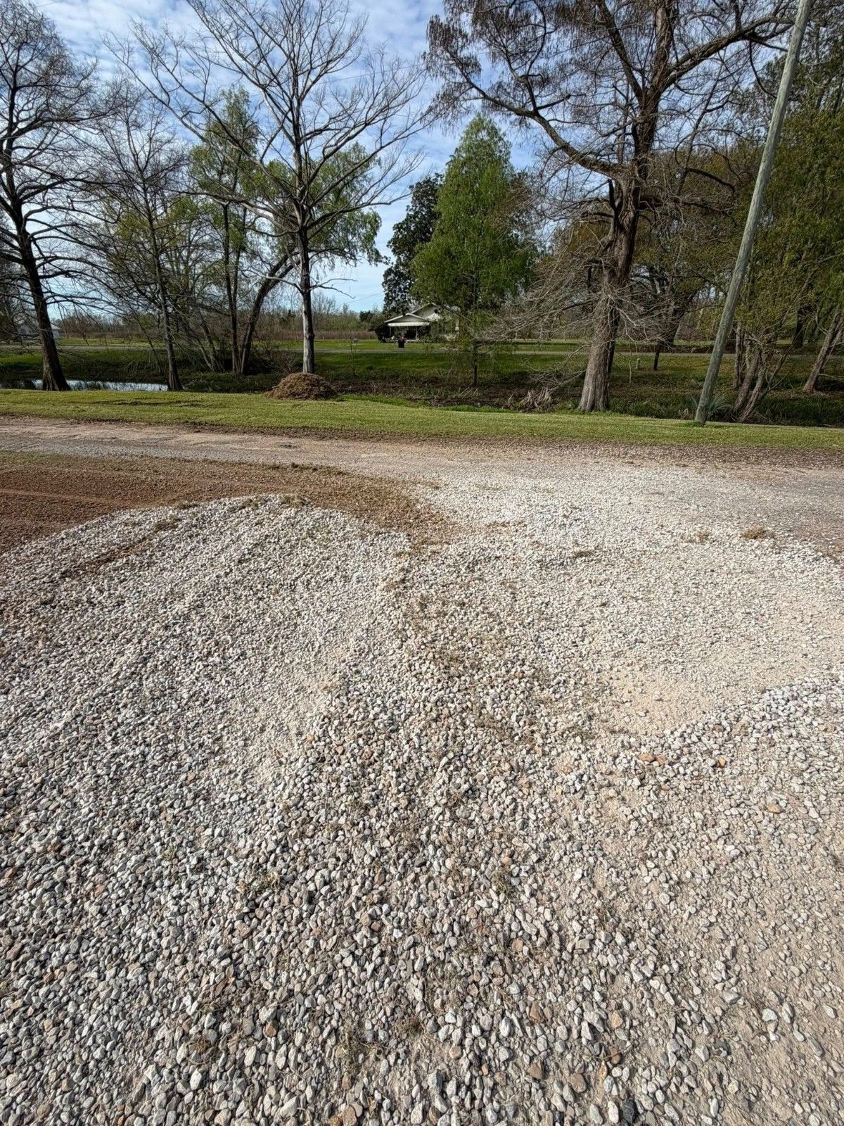 Gravel driveway leading to a grassy area with leafless trees under a partly cloudy sky