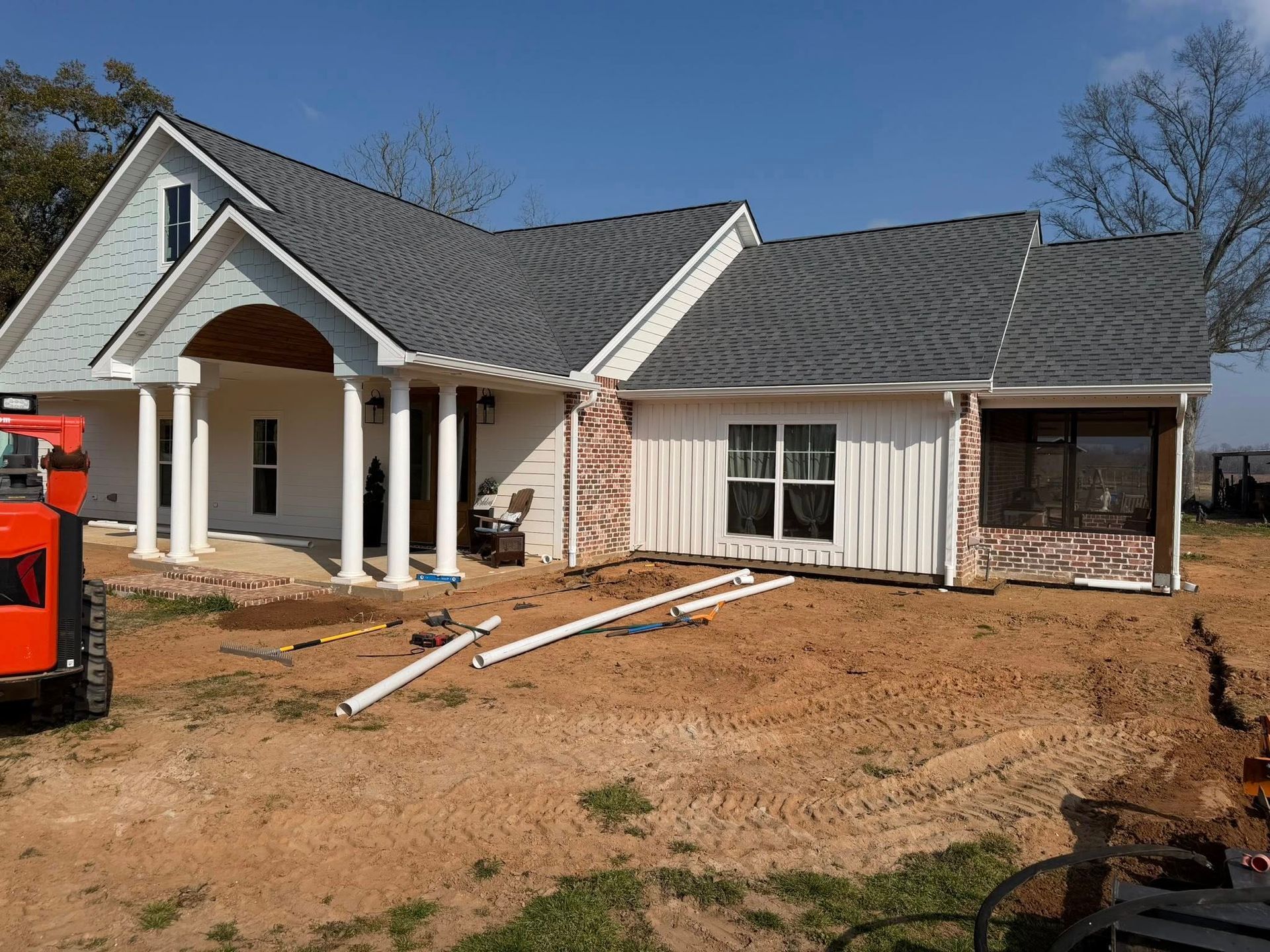 House under construction with front porch, bare dirt yard, and a red construction machine on the left