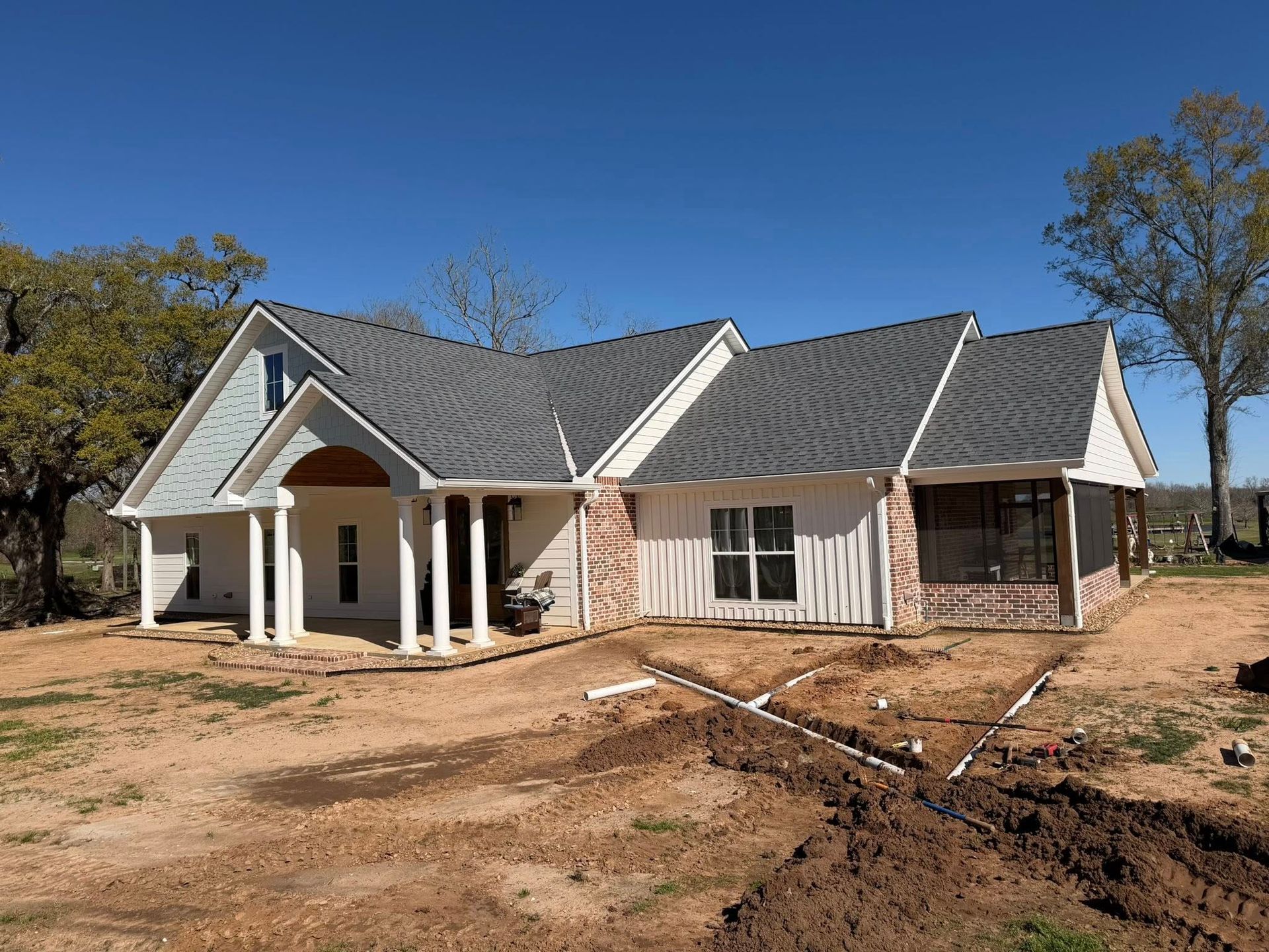 Single-story house with dark gray roof and white trim, surrounded by bare dirt and trees under a blue sky