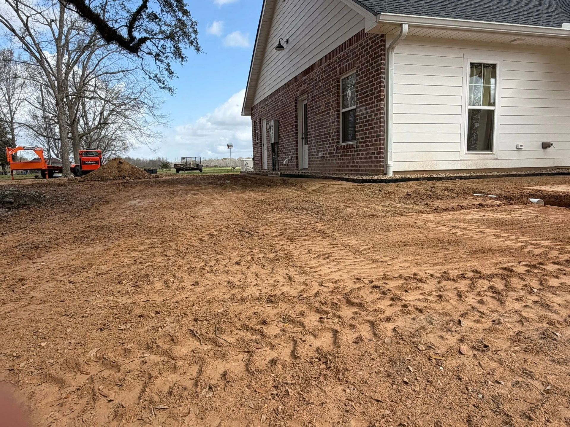 Dirt yard beside a brick-and-white house, with bare trees and two orange construction vehicles in the distance.
