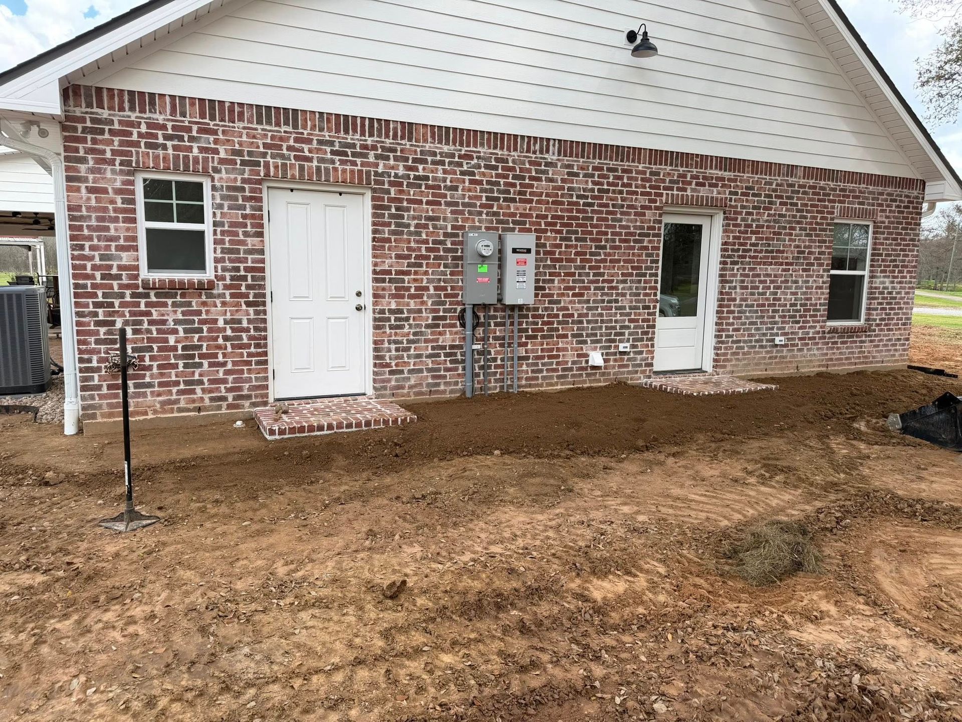 Brick house exterior with white doors, windows, utility meters, and bare dirt yard under construction
