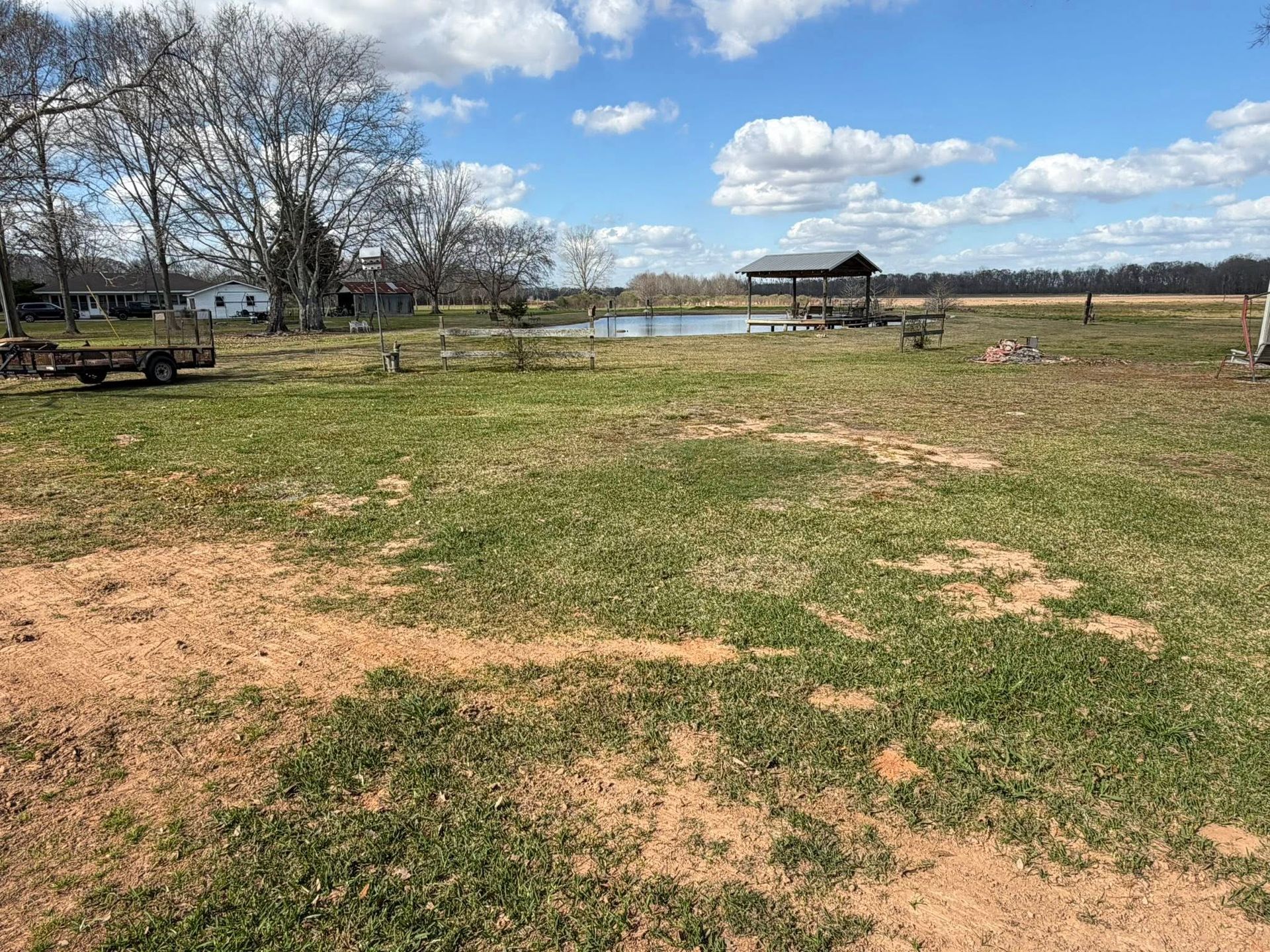 Grassy park by a lake with bare trees, picnic tables, and a pavilion under a blue sky