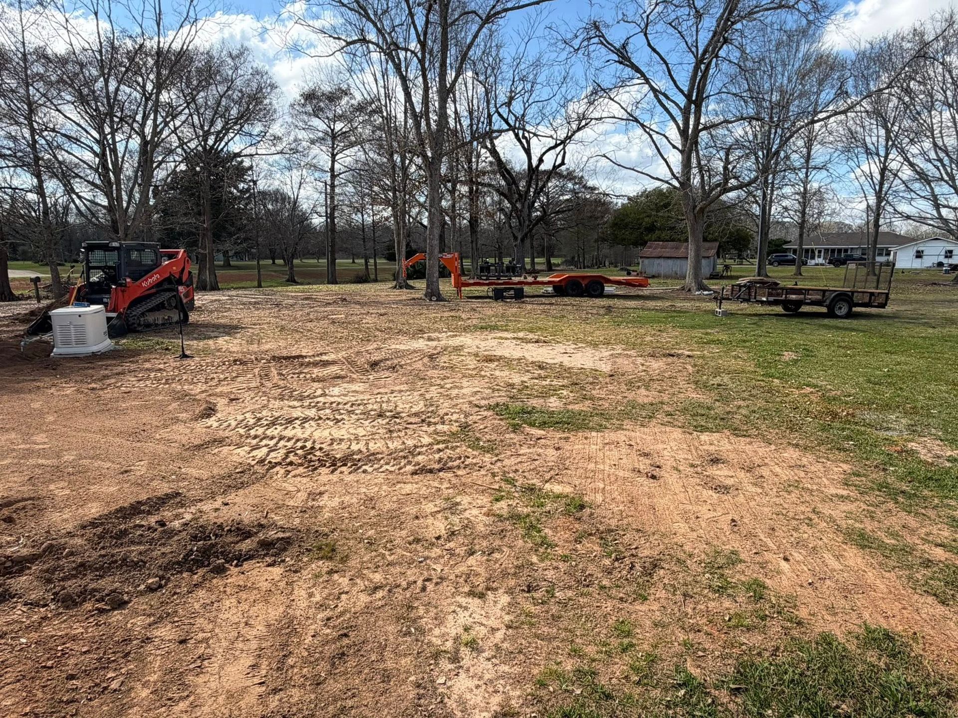 Dirt lot with construction equipment and trailers under leafless trees on a partly cloudy day