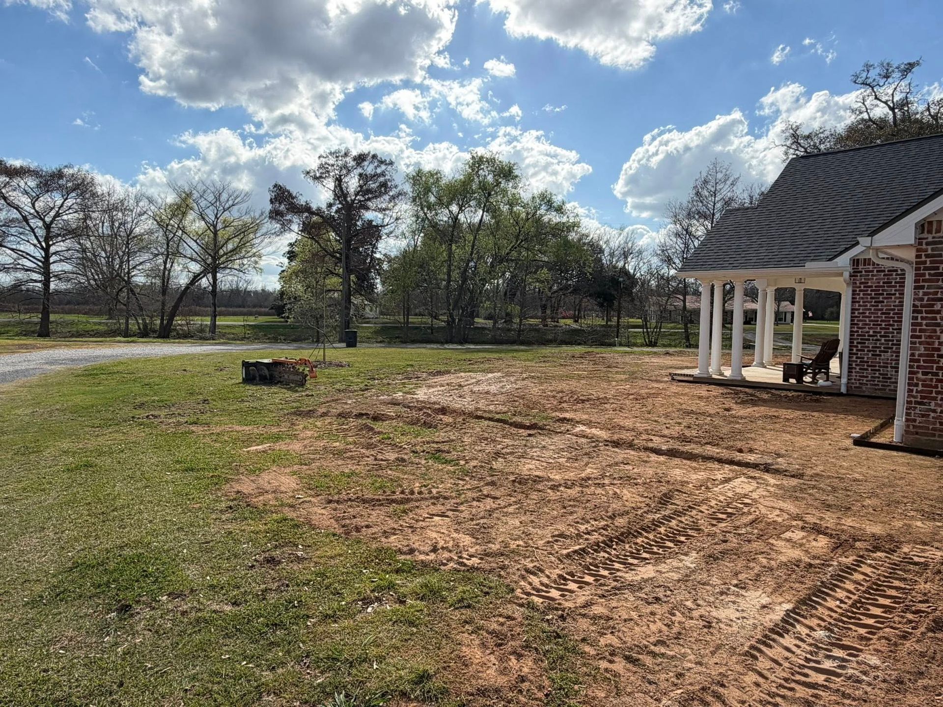 Grassy yard beside a brick house with a porch, under a partly cloudy sky with trees in the distance