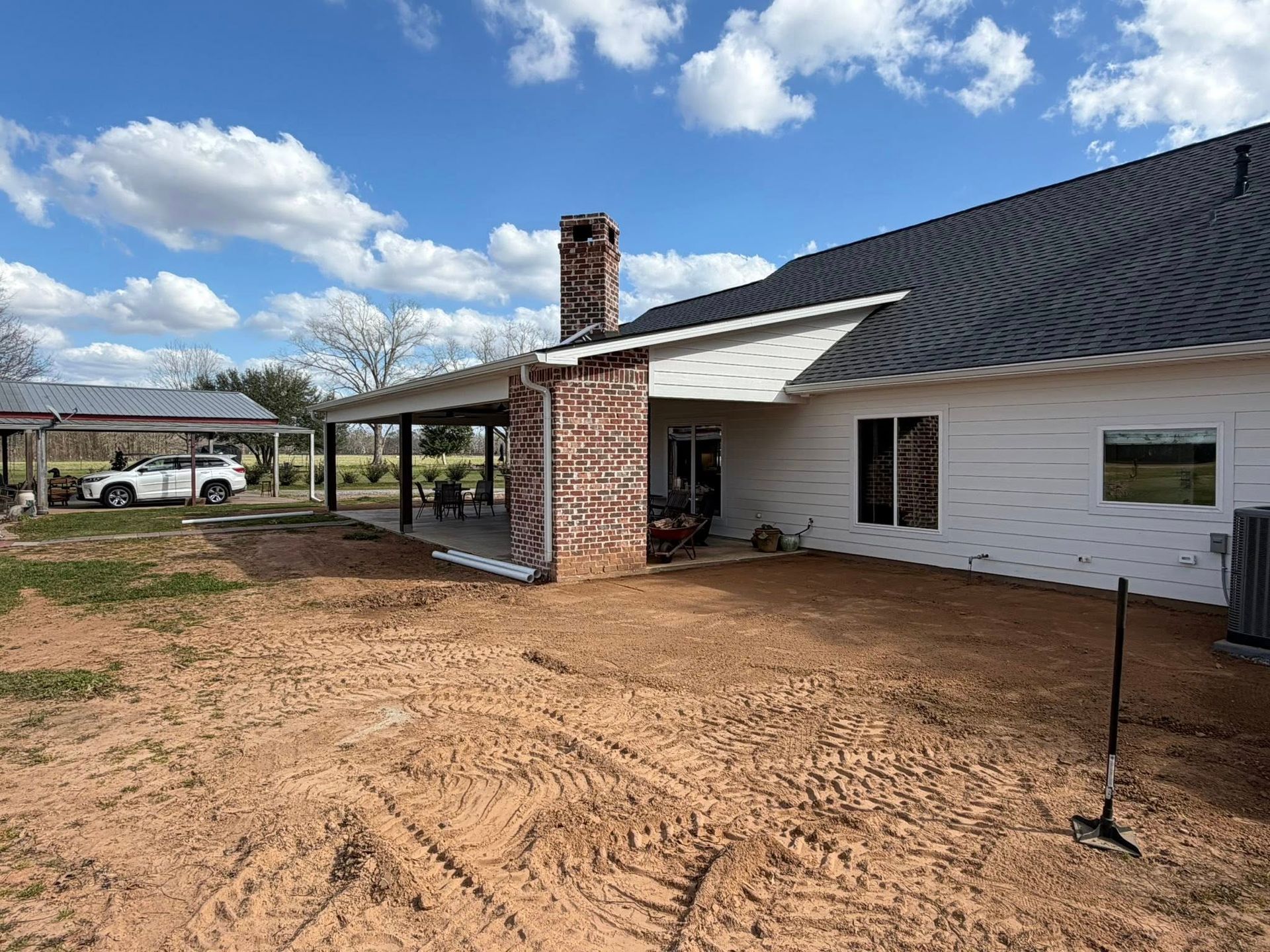 Backyard with a white house, brick chimney, covered patio, and dirt yard under a cloudy sky