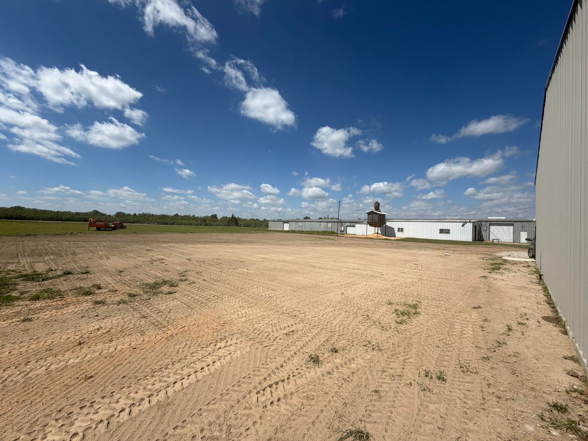 Wide dirt lot beside white industrial buildings under a blue sky with scattered clouds