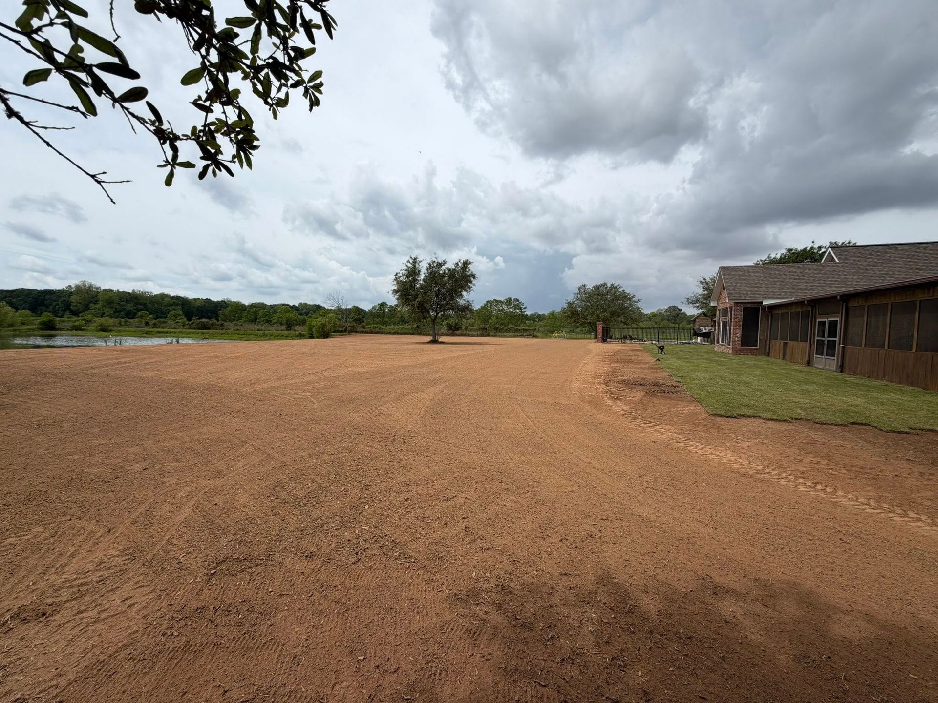 Wide dirt field beside a rural building under cloudy skies, with trees in the distance.