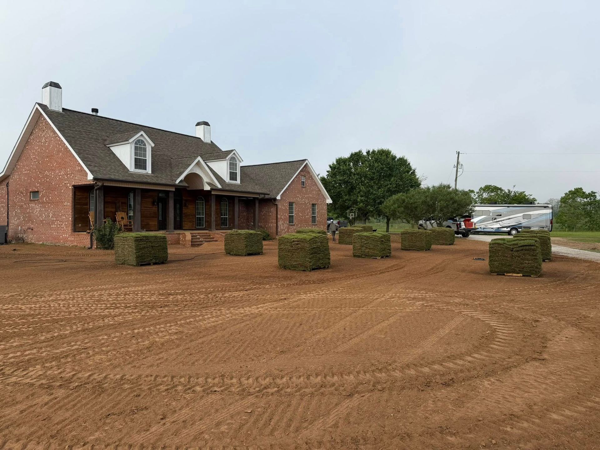 Rural brick house with porch and white-trim roof beside a muddy dirt lot and parked vehicles