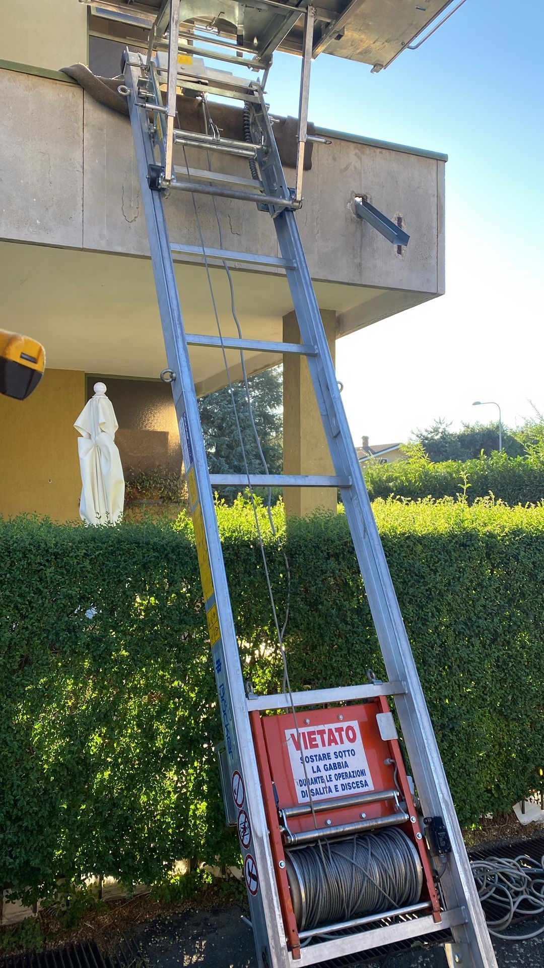Montascale appoggiato a un edificio, utilizzato per spostare oggetti. Edificio beige, siepe verde, giornata di sole.
