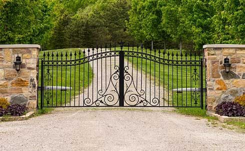 A Wrought Iron Gate Leading To A Gravel Driveway Surrounded By Trees – Mishawaka IN - Milestone Fence