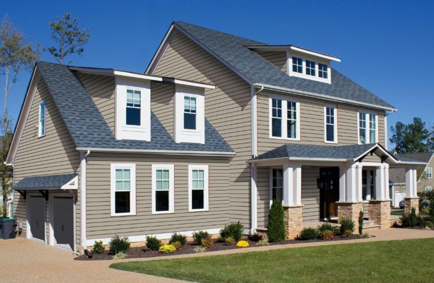 A two-story tan suburban house with a dark shingle roof, two dormer windows, and a front porch on a sunny day.