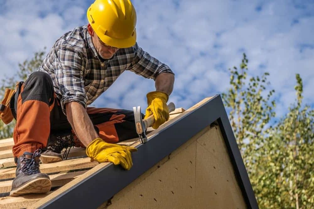 A construction worker in a yellow hard hat and gloves uses a hammer on a roof edge against a blue sky.
