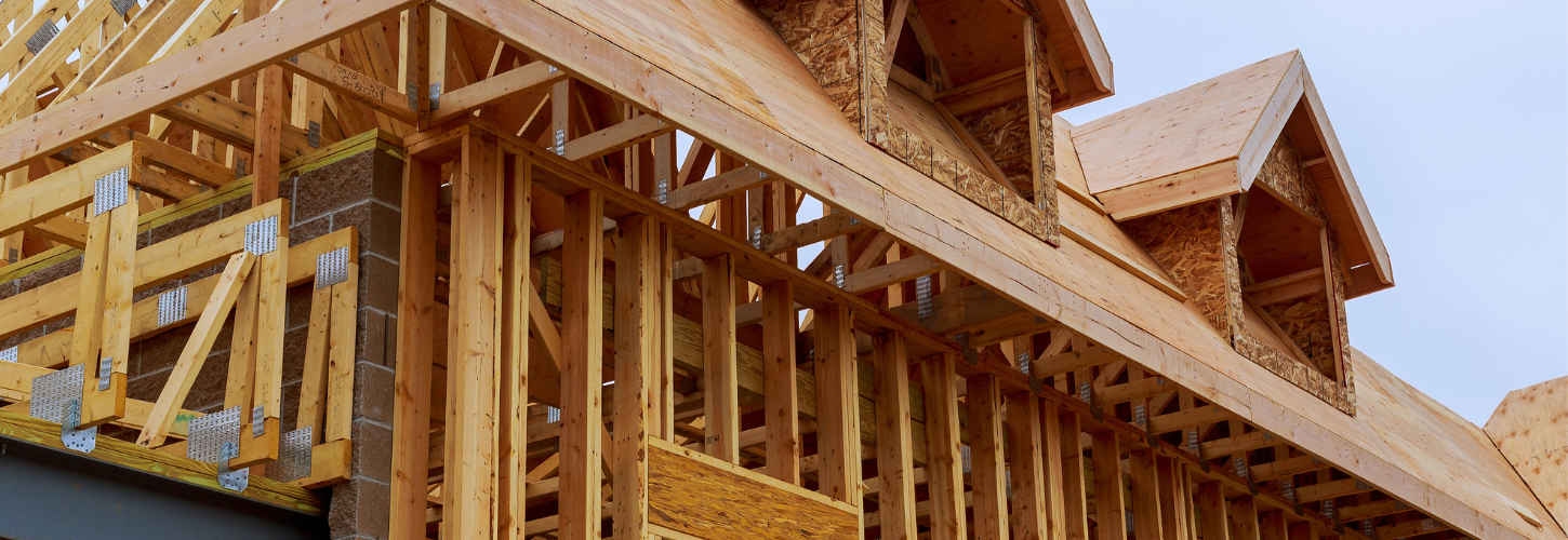The wooden frame of a new house under construction, featuring exposed roof trusses and dormer windows against a clear sky.