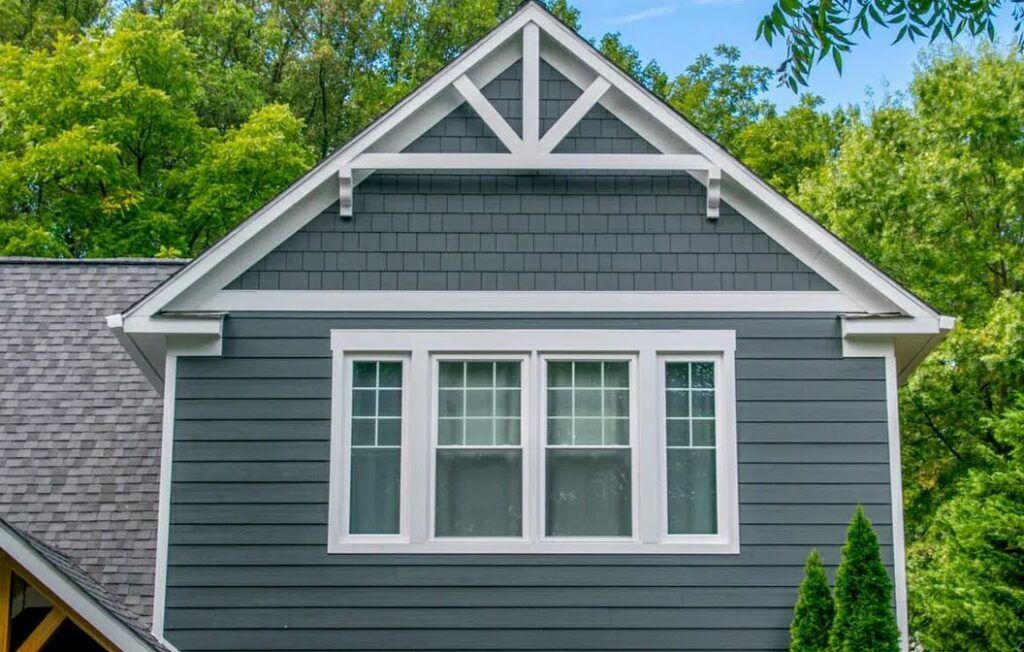 A dark gray-sided house exterior featuring a peaked roof, decorative timber truss, and four white-framed windows.