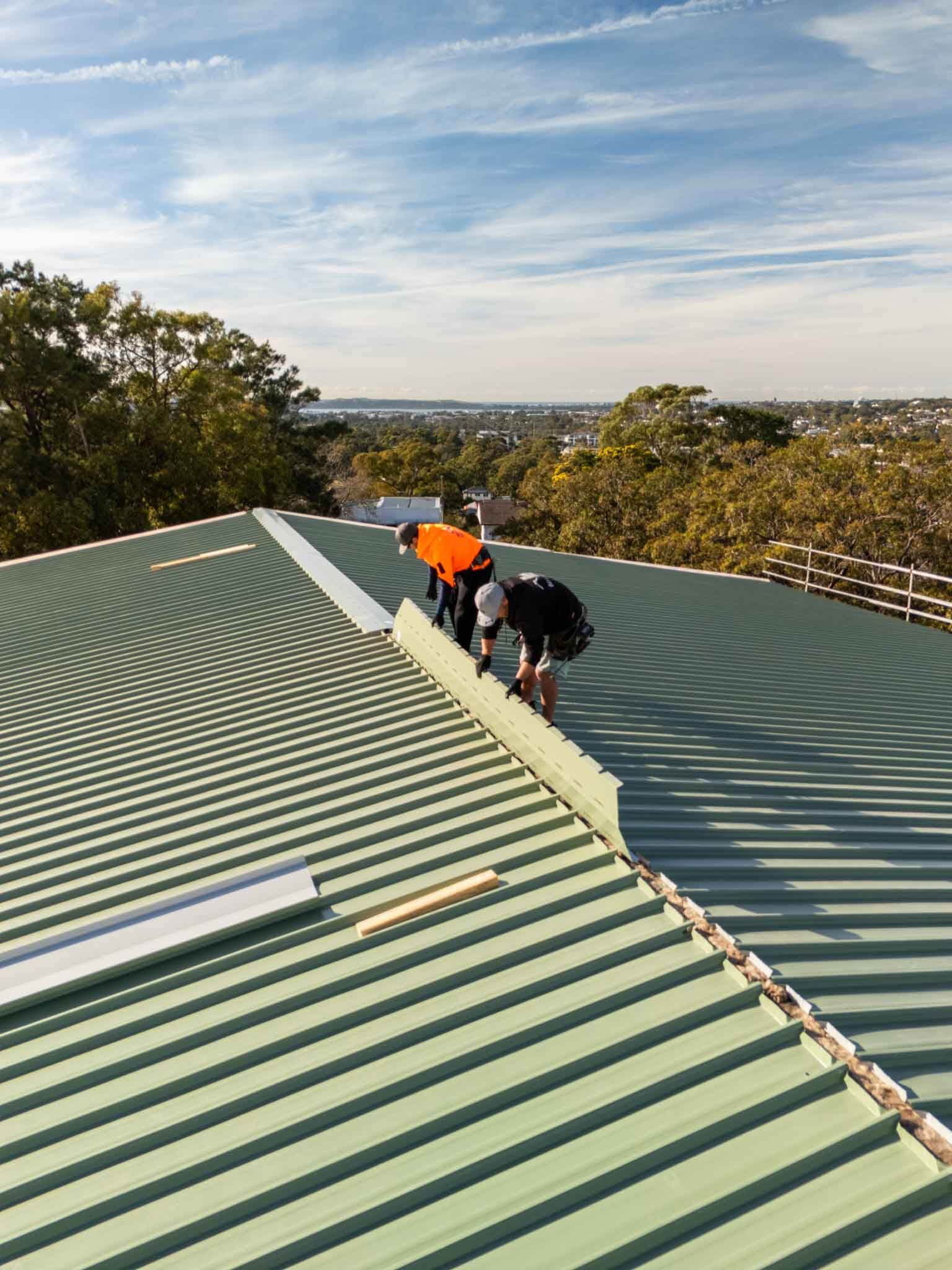 Two workers in high-visibility orange vests installing metal flashing on a green corrugated roof during the day.