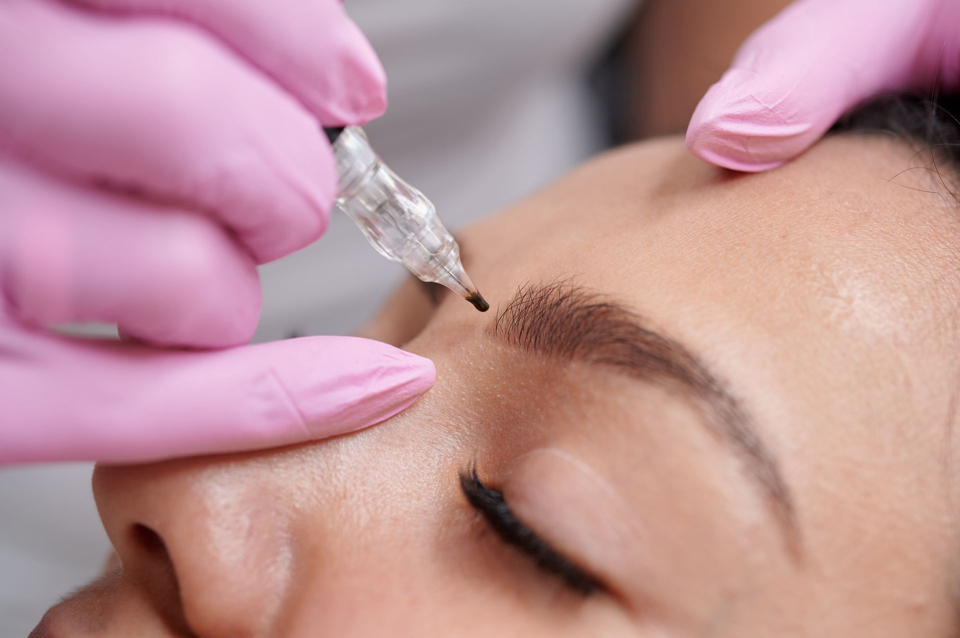 Gloved hands applying cosmetic pigment to an eyebrow with a fine tattooing tool