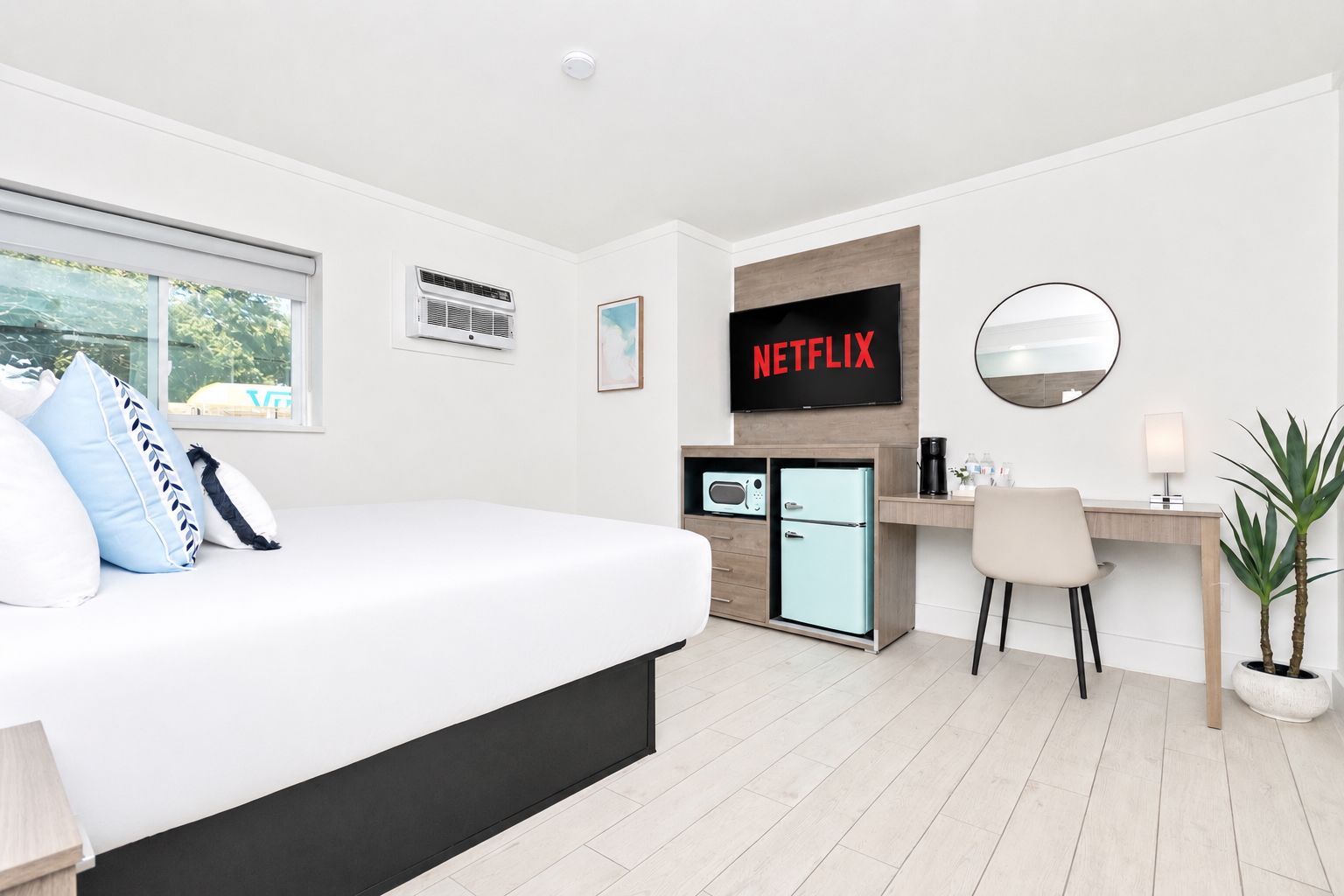 Bedroom with a bed, desk, TV, refrigerator, and window. White walls, wood floor, and neutral decor.