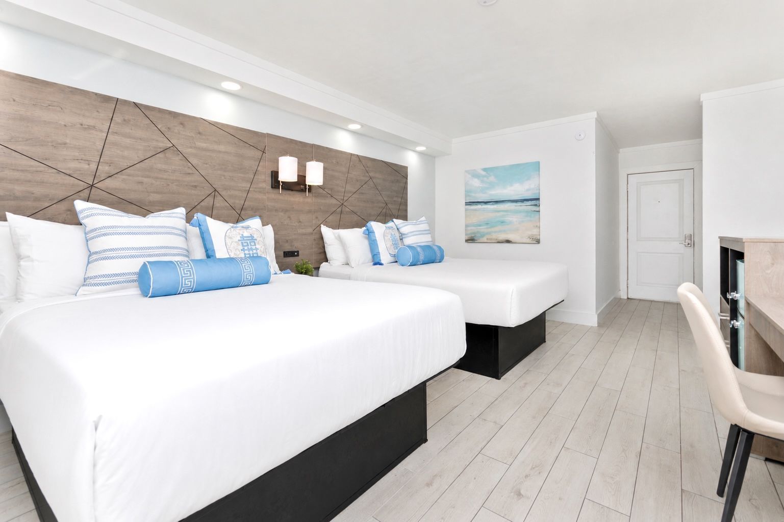Bright modern hotel room with two white beds, blue accents, and light wood flooring