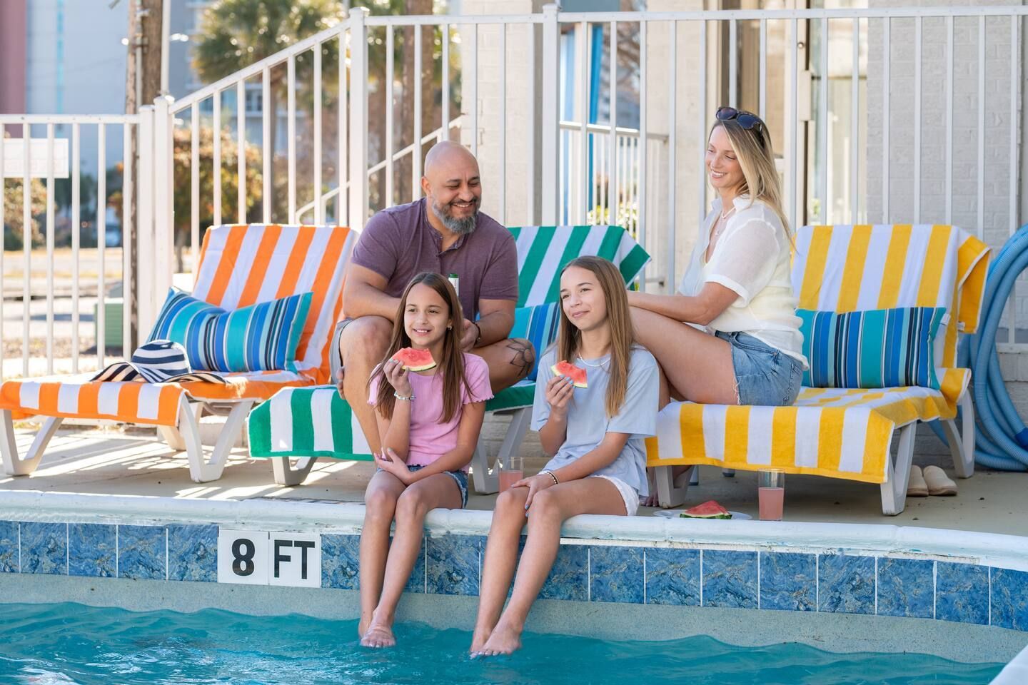 Family sitting by a pool on colorful lounge chairs, feet in the water with an 8 ft depth marker nearby.