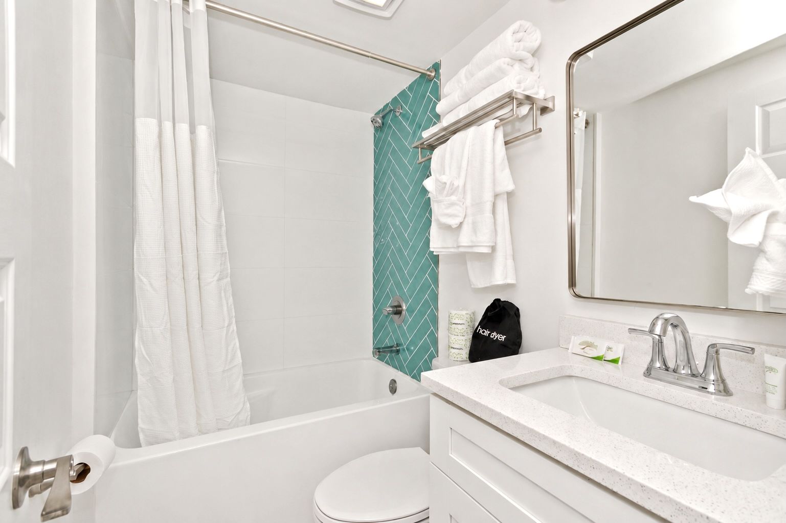 Small white bathroom with bathtub, teal accent tile, vanity sink, and hanging towels.