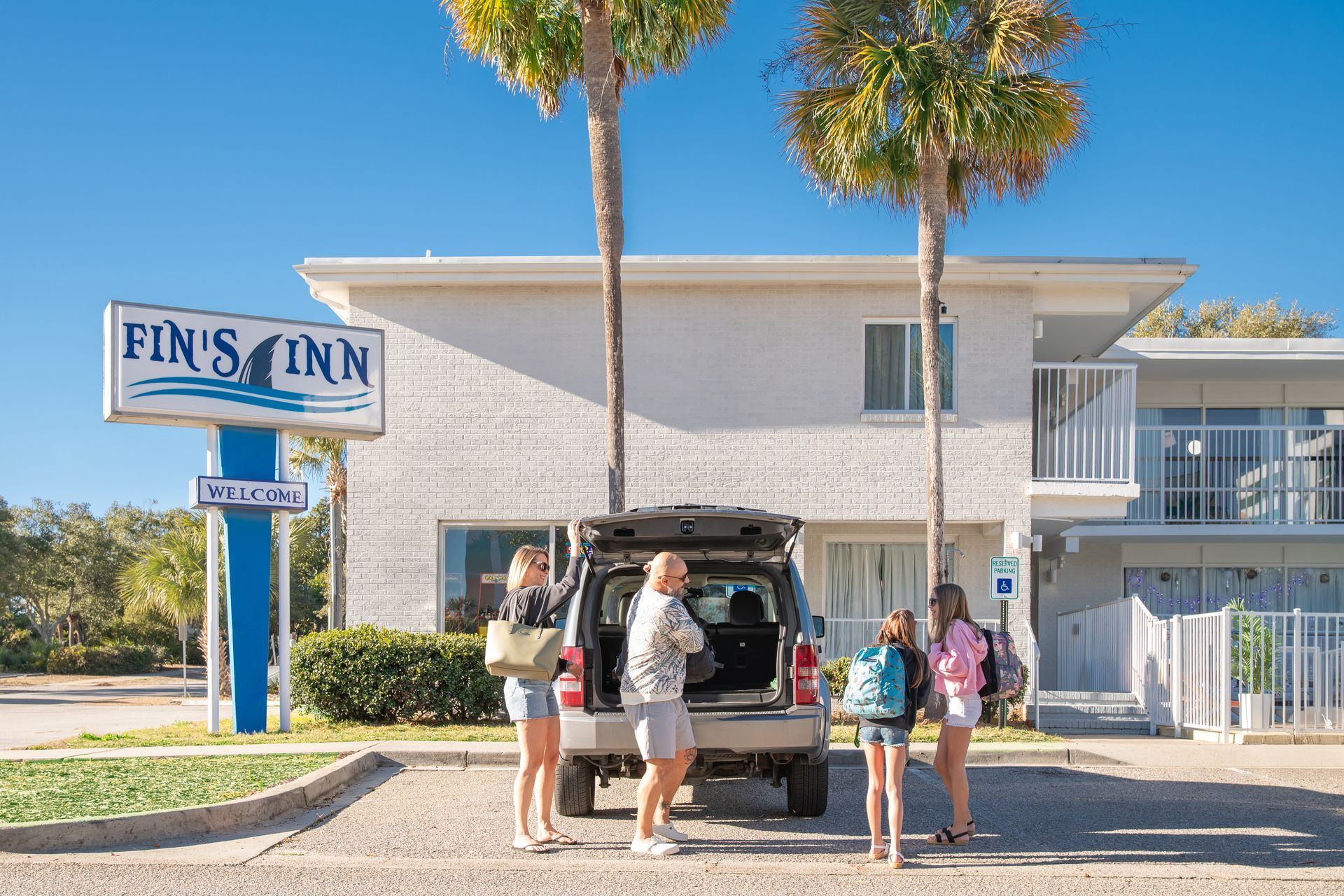 People loading luggage at Fin’s Inn, a white motel with palm trees on a sunny day