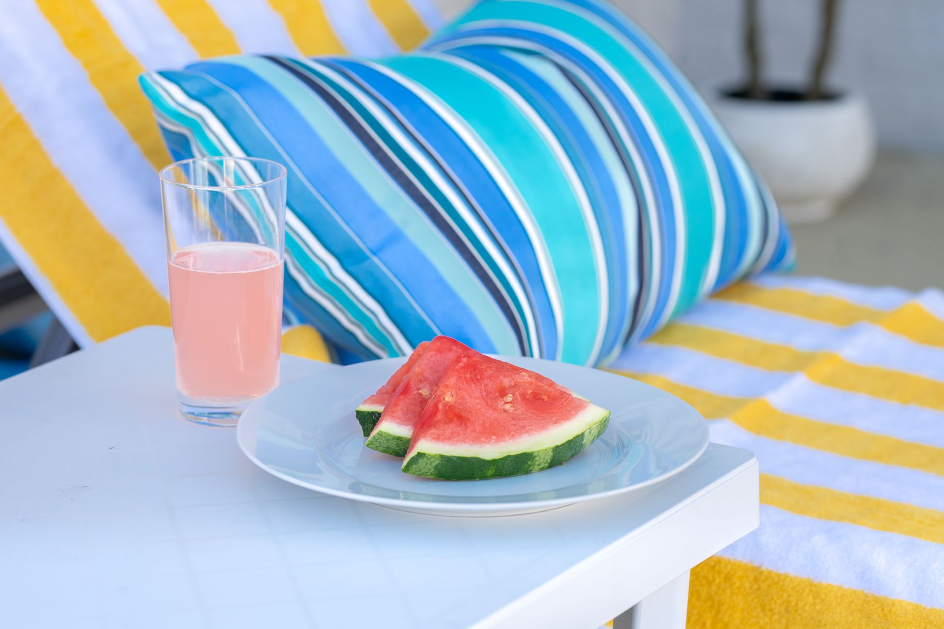 Pink drink and watermelon slices on a tray beside striped cushions at a beachside lounge chair
