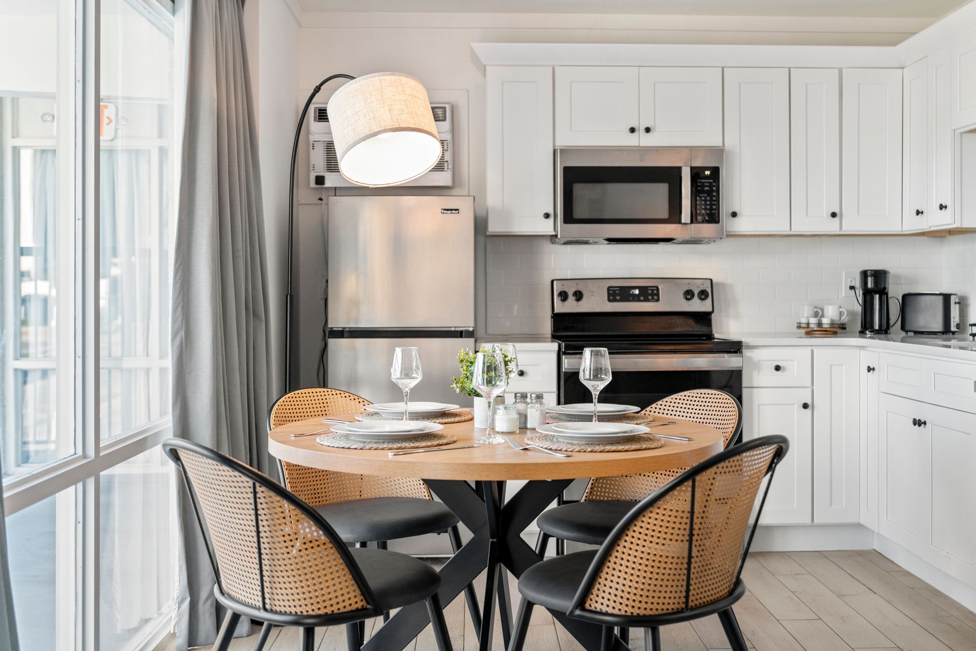 Bright white kitchen with a round dining table, black chairs, stainless appliances, and large windows.