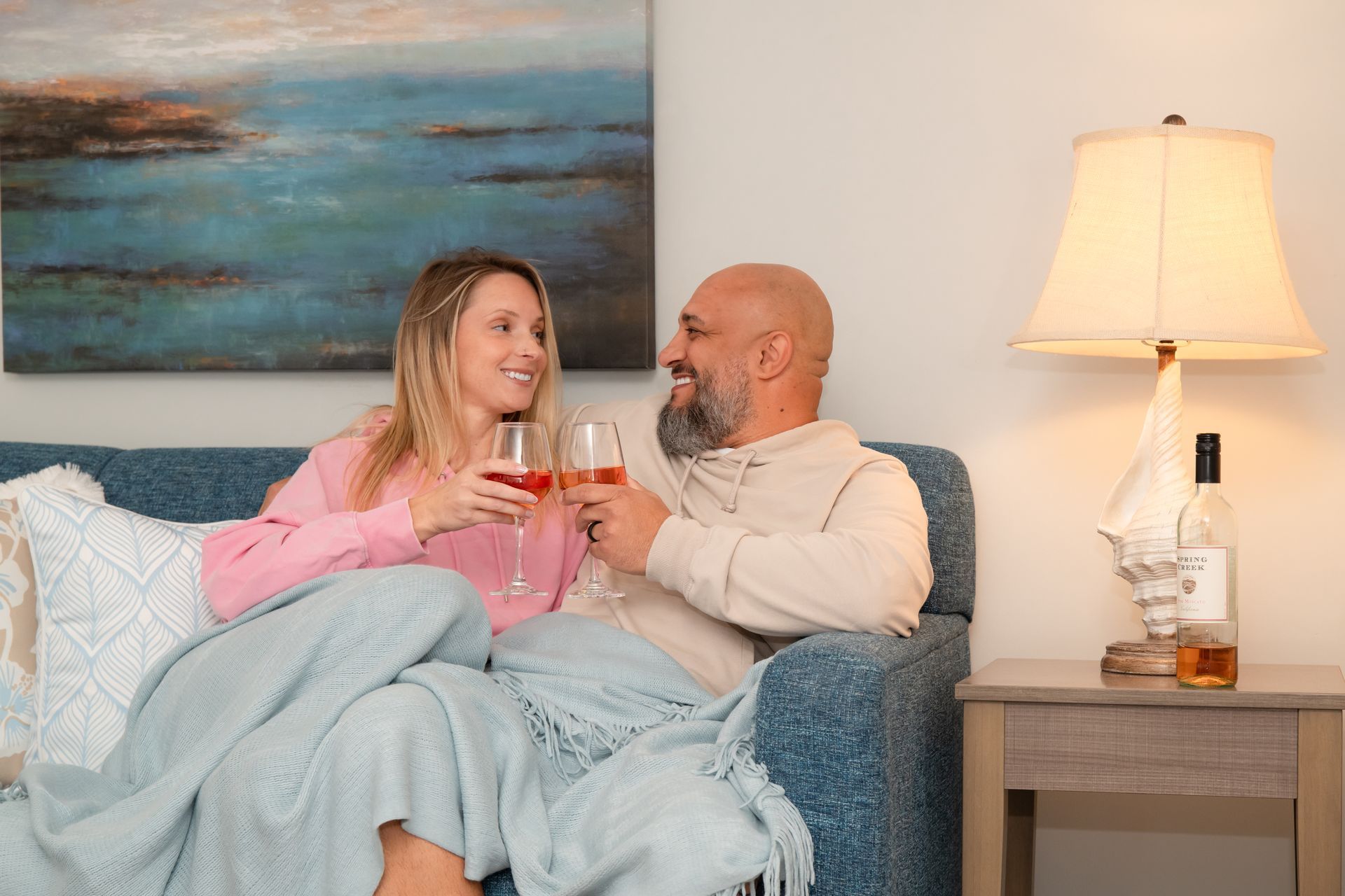 Couple on a couch toasting drinks in a cozy living room with a lamp and wall art.