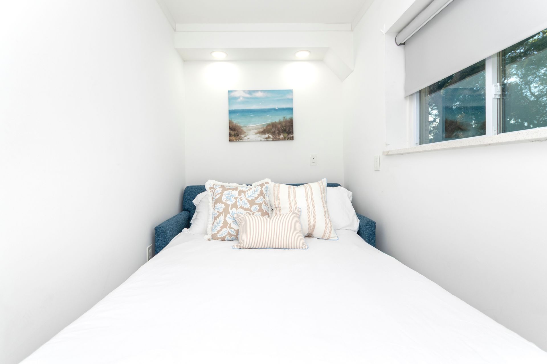 Narrow white bedroom with a bed, patterned pillows, wall art, and a small window