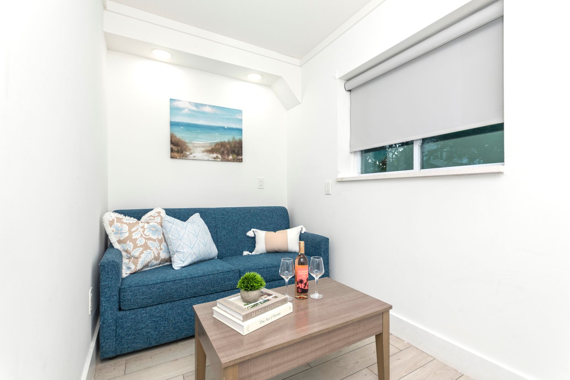 Bright living room with a blue sofa, coffee table, wall art, and a window with a blind