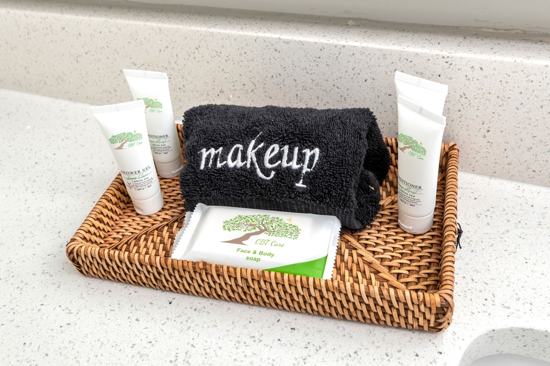 Bathroom toiletries arranged on a woven tray. Includes tubes of lotion, soap, and a makeup washcloth with a small plant.