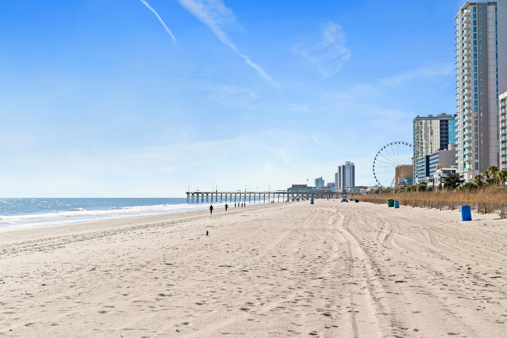 Sunny beach with sandy shore, blue ocean, boardwalk, and distant high-rise buildings under a clear sky
