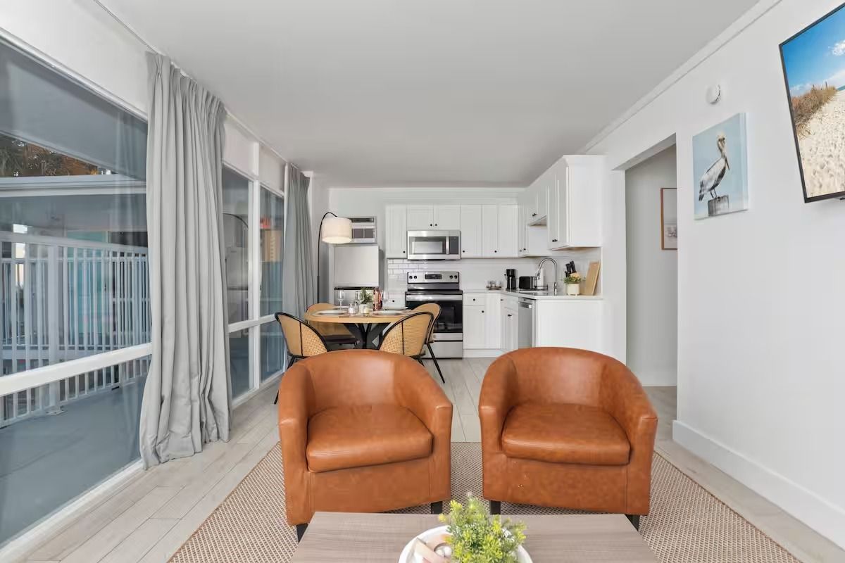 Bright living room with two tan chairs, a dining table, and a white kitchen opening to a balcony.