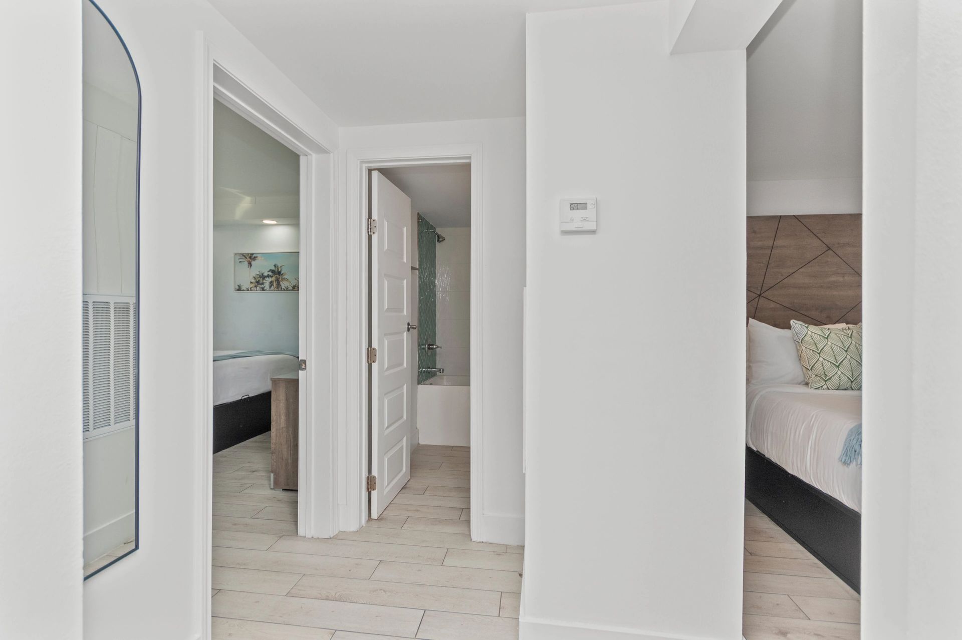 Bright white hallway with open doorways to a bedroom and bathroom, featuring light wood floors and a bed visible.