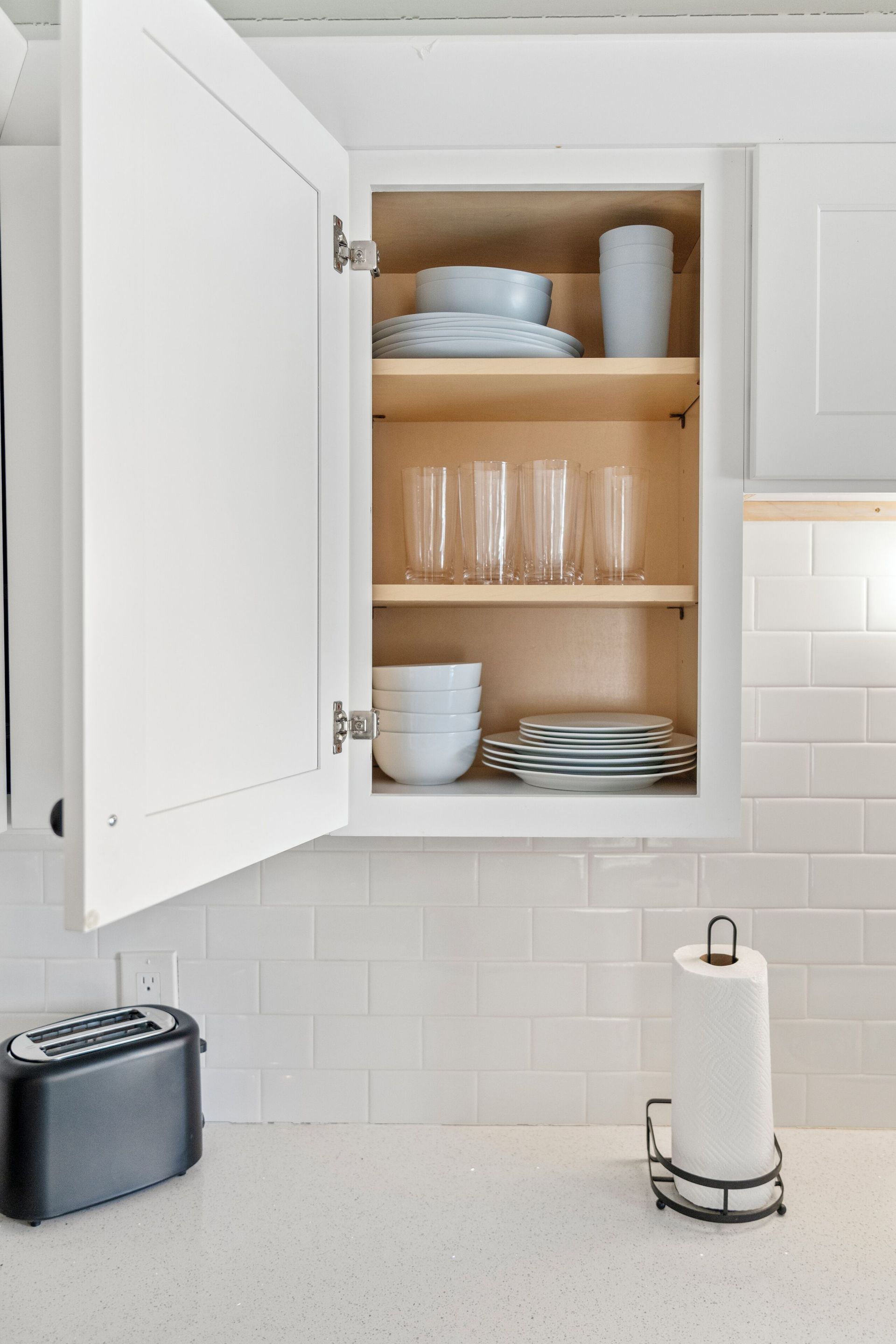 Open kitchen cabinet with dishes, glasses, and mugs on shelves beside a white tiled backsplash.