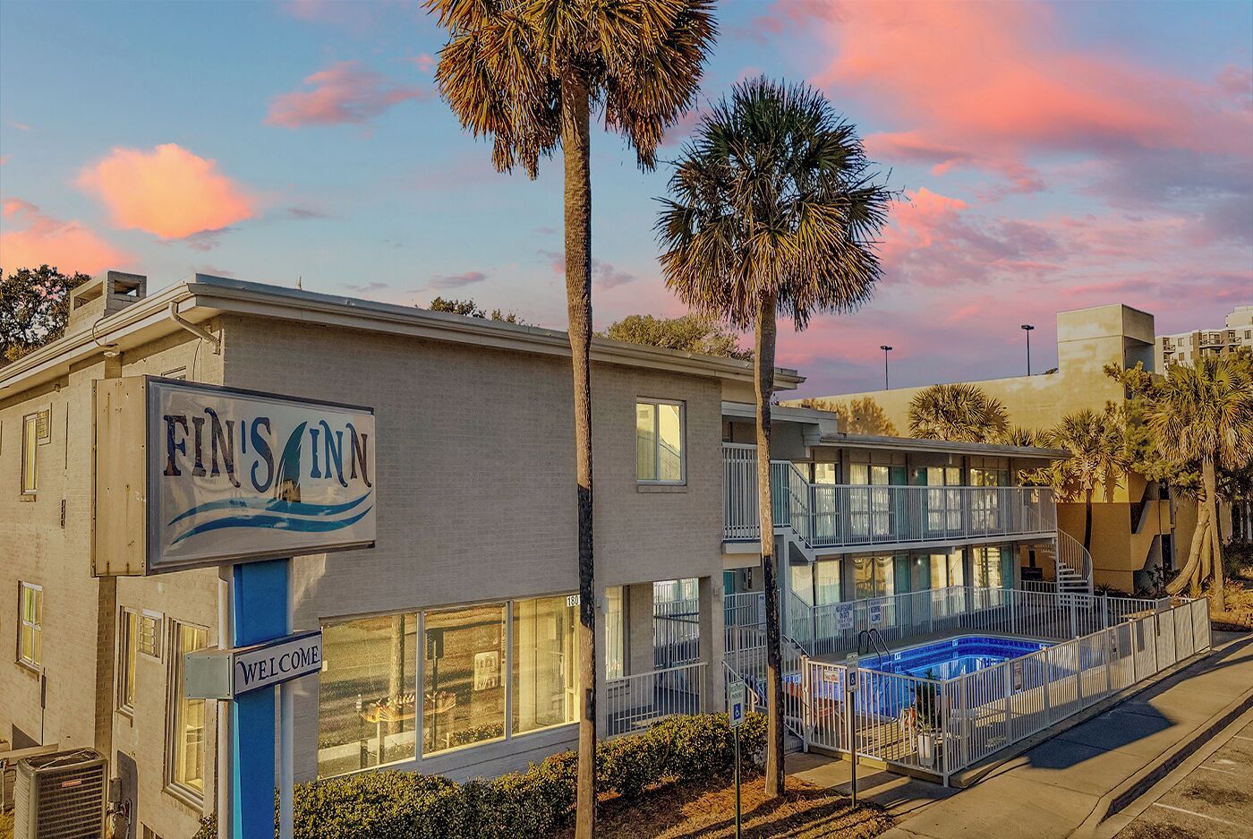 Sunset view of a beachside motel with palm trees, balconies, and a blue pool under a pink sky