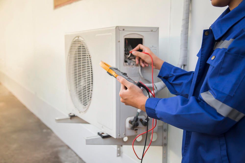 HVAC technician in blue overalls tests an air conditioner unit with a multimeter.