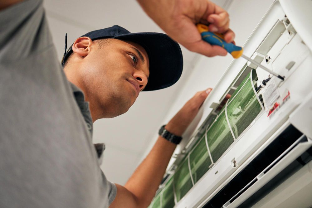 HVAC technician fixing an air conditioner with a screwdriver. Inside a building, wearing a cap.