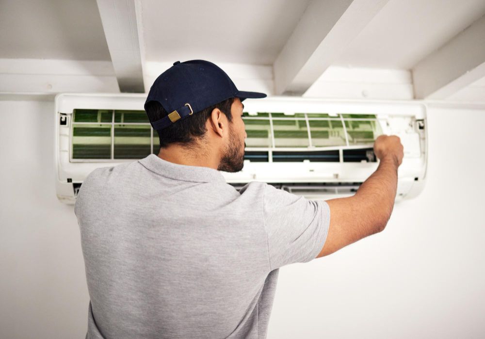 Person in cap cleaning an air conditioner filter on a white wall.