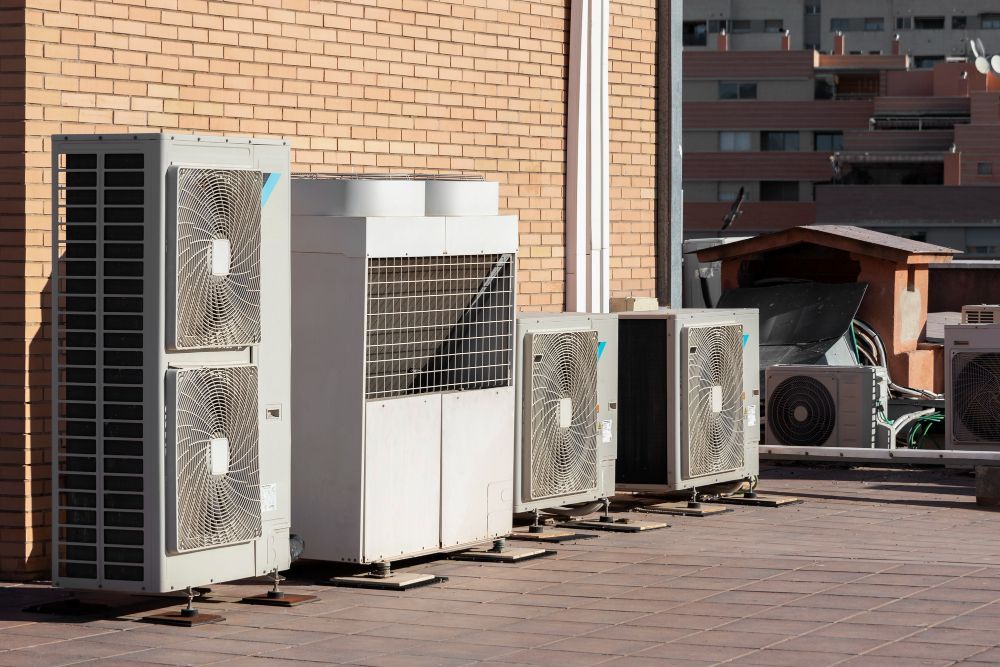 Air conditioning units line a brick wall on a rooftop, with a city skyline in the background.