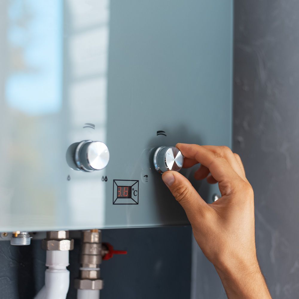 Hand turning a dial on a grey water heater. White pipes and a digital temperature display are visible.