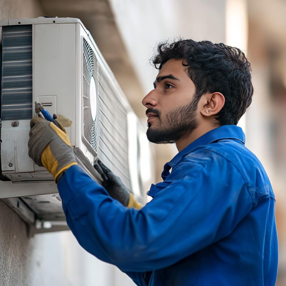 HVAC technician in blue jumpsuit, inspecting an AC unit outdoors, using a tool.