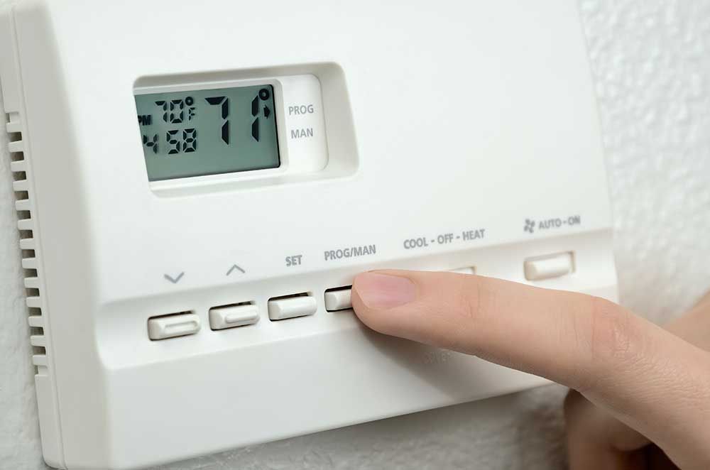 Hand pressing the “program” button on a white digital thermostat, displaying 70°F.