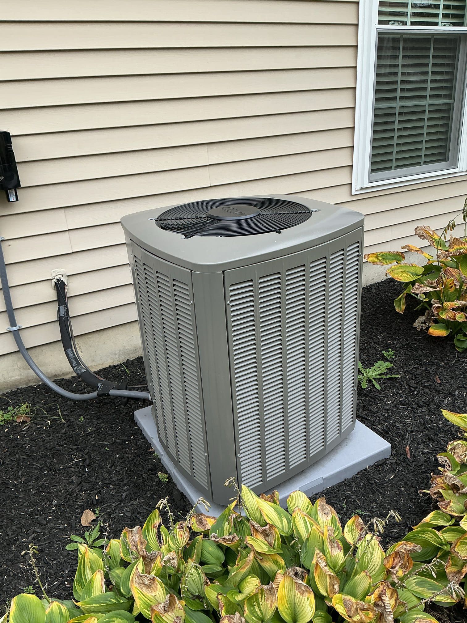 Gray air conditioning unit outside a beige house with a window and shrubs.