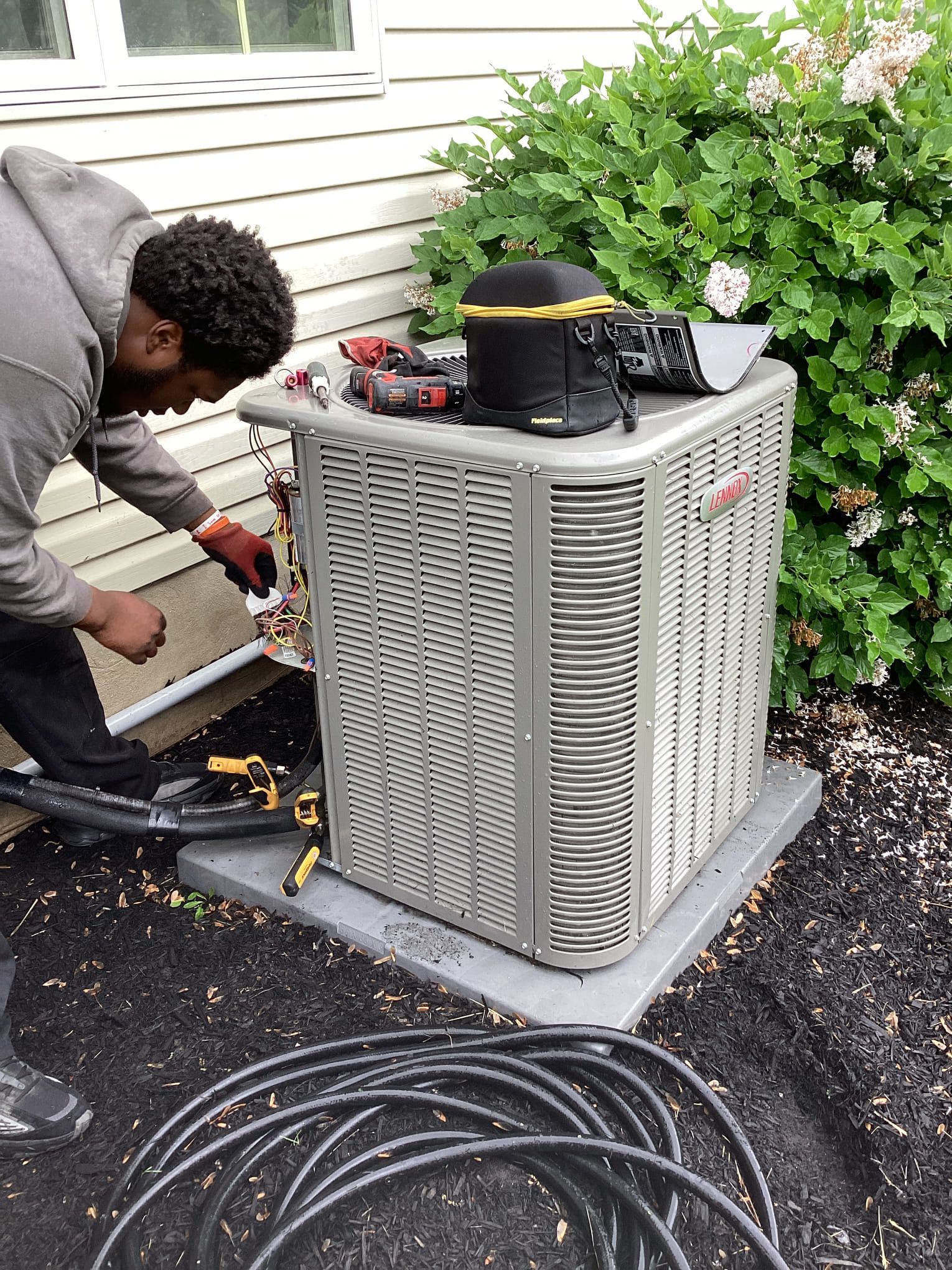 HVAC technician working on an outdoor air conditioning unit, next to a bush, tools on top.