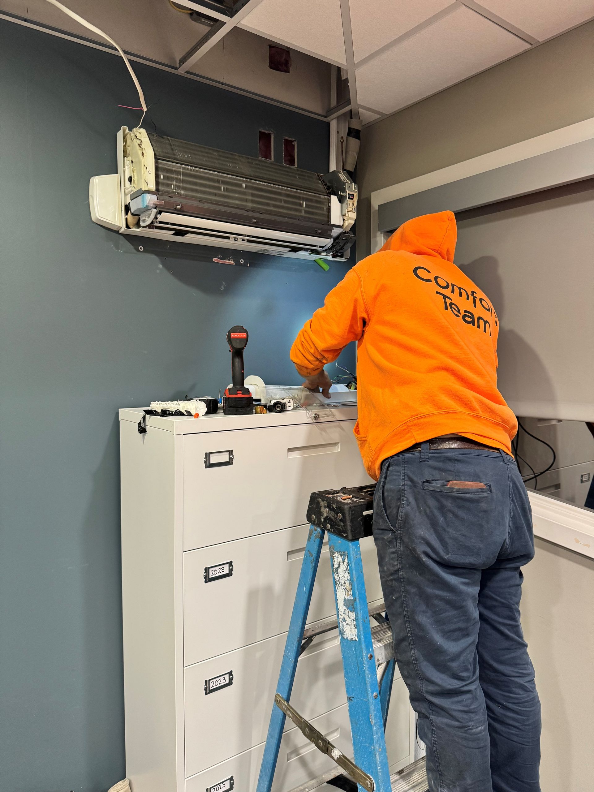 A person in an orange hoodie repairs an AC unit on a blue wall, standing on a ladder next to a file cabinet.