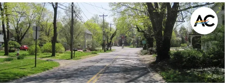 Street lined with trees, houses, and power lines. A cyclist rides in the distance.