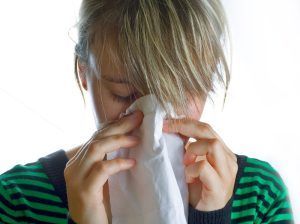 Woman blowing her nose with a white tissue; wearing a green and black striped shirt.