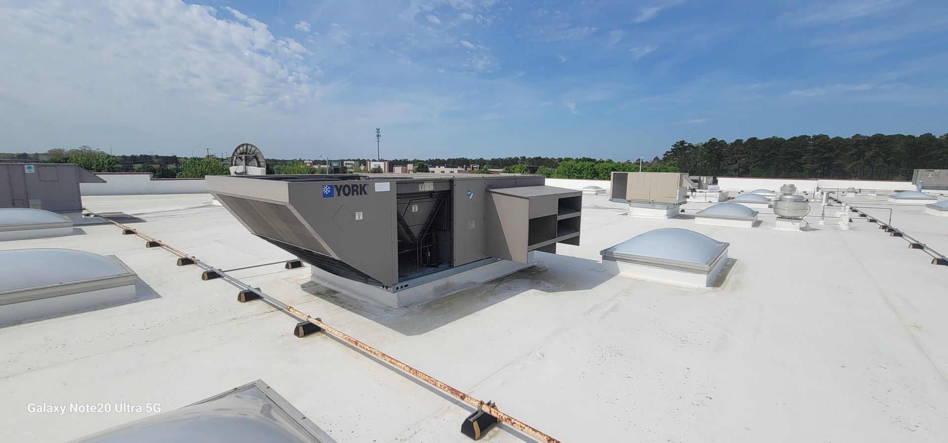 HVAC unit on a white commercial roof under a blue sky with some trees in the background.