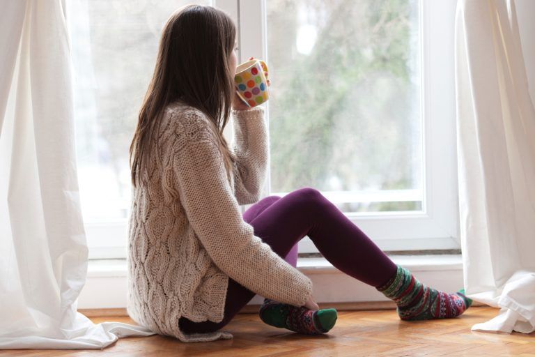 Woman sitting by window, sipping from colorful mug; wearing sweater, leggings, and patterned socks.